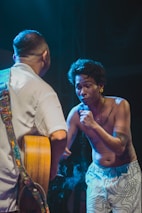 Two musicians are captured in an intense moment on stage. One is shirtless with tattoos, passionately singing or speaking into a microphone, while the other wears a light-colored shirt and plays a guitar. Smoke is visible, adding to the dramatic atmosphere.