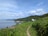 A coastal landscape surrounded by lush green grass and trees, leading down a path to a small white house nestled on a hill. The ocean is calm and stretches to the horizon under a blue sky with scattered clouds.