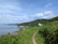 A coastal landscape surrounded by lush green grass and trees, leading down a path to a small white house nestled on a hill. The ocean is calm and stretches to the horizon under a blue sky with scattered clouds.