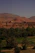A scenic view of a desert landscape with an oasis. In the foreground, lush palm trees and cultivated green fields provide a stark contrast to the arid environment. In the background, a small village composed of traditional clay buildings sits at the base of a dramatic rocky hill with expansive mountains under a clear blue sky.