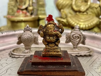 A small bronze figurine of a deity adorned with red powder is placed on a wooden platform. Surrounding the central figure are ornate metallic designs, possibly depicting other religious symbols or icons. The background has additional details with similar metallic textures and designs.