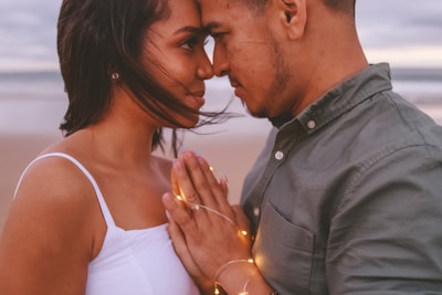 A couple stands close together, facing each other with their foreheads touching. The woman's hair is gently blowing in the wind, and they are holding hands wrapped with small string lights. The background shows a blurred beach setting, suggesting an intimate moment by the sea.