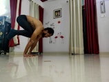 A person balances in a yoga pose on a shiny floor in a room. The background features decorative elements such as flamingo wall art, framed pictures, and contrasting red and cream curtains.