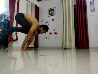 A person balances in a yoga pose on a shiny floor in a room. The background features decorative elements such as flamingo wall art, framed pictures, and contrasting red and cream curtains.