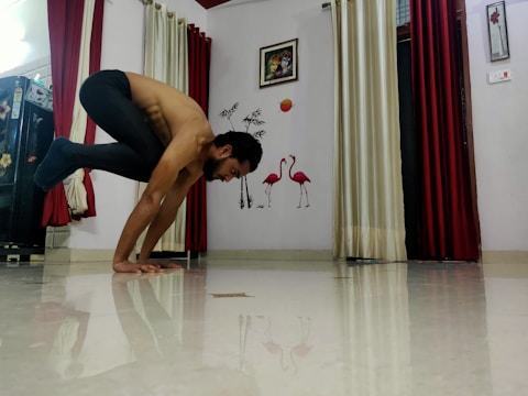 A person balances in a yoga pose on a shiny floor in a room. The background features decorative elements such as flamingo wall art, framed pictures, and contrasting red and cream curtains.