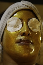 A person is lying down with a radiant gold facial mask applied to their face, covering the skin entirely except for their mouth. Both eyes are covered with circular cotton pads, and a white headband is wrapped around the forehead, holding the hair back.