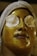 A person is lying down with a radiant gold facial mask applied to their face, covering the skin entirely except for their mouth. Both eyes are covered with circular cotton pads, and a white headband is wrapped around the forehead, holding the hair back.