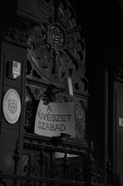 An ornate, arched doorway with intricate carvings and a central circular emblem. A sign with the text 'A MŰVÉSZET SZABAD' is hung over the decorative railing. The architecture is detailed with symmetrical patterns and motifs.