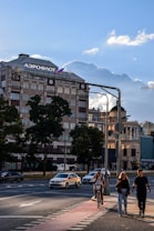 A city scene depicting a large building with the word 'Аэрофлот' on top and a Russian flag. Several people are walking on the sidewalk, one of them is riding a scooter. There are trees, streetlights, and vehicles including cars and taxis on the road. The weather appears clear with a blue sky and some clouds.