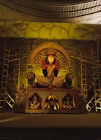 A grand idol of a multi-armed deity, possibly Durga, is adorned with intricate details. The statue is surrounded by lions and other figures, set in a dimly lit environment with decorative scaffolding and elaborate patterns on the walls.