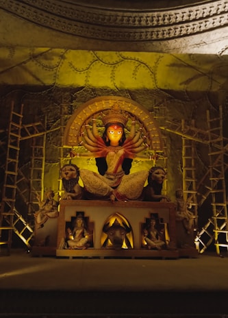A grand idol of a multi-armed deity, possibly Durga, is adorned with intricate details. The statue is surrounded by lions and other figures, set in a dimly lit environment with decorative scaffolding and elaborate patterns on the walls.