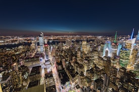 A stunning aerial view of a cityscape at night, featuring an array of illuminated skyscrapers and vibrant lights. The skyline includes modern high-rise buildings adorned with colorful lights, while the background shows a dark sky transitioning to dusk, with a river cutting through the city.