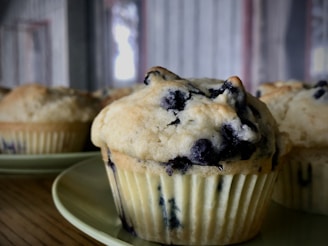 A close-up of a blueberry muffin with visible blueberries embedded in the golden brown, textured surface. The muffin sits on a pale green plate with a blurred background featuring additional muffins and soft lighting.