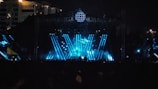 A large outdoor concert stage with vibrant blue and white lights illuminating the scene. The stage structure is enhanced by beams of light forming dynamic patterns, creating a visually striking atmosphere. The silhouette of a performer is visible at the center, positioned behind a DJ table or similar setup. The crowd is mostly in shadow, emphasizing the energetic lighting on the stage. In the background, a city skyline is partially visible, with some illuminated buildings.