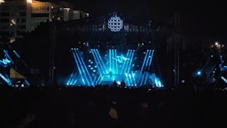 A large outdoor concert stage with vibrant blue and white lights illuminating the scene. The stage structure is enhanced by beams of light forming dynamic patterns, creating a visually striking atmosphere. The silhouette of a performer is visible at the center, positioned behind a DJ table or similar setup. The crowd is mostly in shadow, emphasizing the energetic lighting on the stage. In the background, a city skyline is partially visible, with some illuminated buildings.