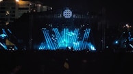 A large outdoor concert stage with vibrant blue and white lights illuminating the scene. The stage structure is enhanced by beams of light forming dynamic patterns, creating a visually striking atmosphere. The silhouette of a performer is visible at the center, positioned behind a DJ table or similar setup. The crowd is mostly in shadow, emphasizing the energetic lighting on the stage. In the background, a city skyline is partially visible, with some illuminated buildings.
