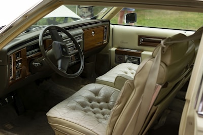 The interior of a vintage car features beige leather seats with a plush, tufted design. Wood paneling accents the dashboard, which also contains classic analog controls and a large steering wheel. The interior appears well-maintained, emanating a sense of luxury from a past era.