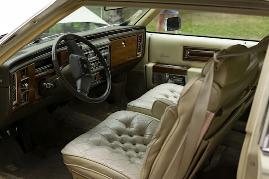 The interior of a vintage car features beige leather seats with a plush, tufted design. Wood paneling accents the dashboard, which also contains classic analog controls and a large steering wheel. The interior appears well-maintained, emanating a sense of luxury from a past era.
