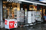 A street-side souvenir shop displaying various items for sale. Outside the shop, there are rotating stands filled with postcards, magnets, and other small souvenirs. An ice cream freezer with the brand 'OLA' is visible in front, featuring images of strawberries and chocolate. The store's interior is well-lit, showcasing an array of products on shelves.