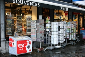 A street-side souvenir shop displaying various items for sale. Outside the shop, there are rotating stands filled with postcards, magnets, and other small souvenirs. An ice cream freezer with the brand 'OLA' is visible in front, featuring images of strawberries and chocolate. The store's interior is well-lit, showcasing an array of products on shelves.