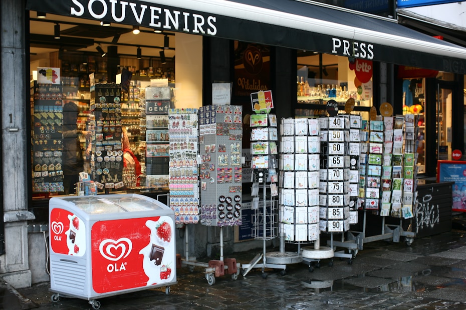 A street-side souvenir shop displaying various items for sale. Outside the shop, there are rotating stands filled with postcards, magnets, and other small souvenirs. An ice cream freezer with the brand 'OLA' is visible in front, featuring images of strawberries and chocolate. The store's interior is well-lit, showcasing an array of products on shelves.