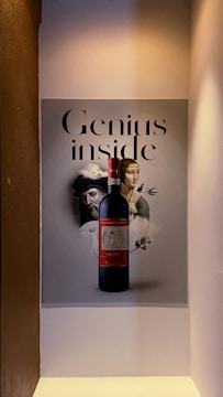 A bottle of red wine is placed in front of a poster that features the words 'Genius inside'. The poster also has images of classical artwork, including a portrait of a man in a turban, a woman, and a sketch of a flying machine.