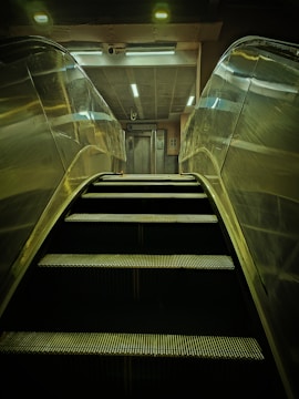 An escalator is ascending towards a well-lit area featuring an elevator. The scene is mostly metallic with reflections on the escalator sides, and overhead lighting provides a dim glow.