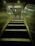 An escalator is ascending towards a well-lit area featuring an elevator. The scene is mostly metallic with reflections on the escalator sides, and overhead lighting provides a dim glow.