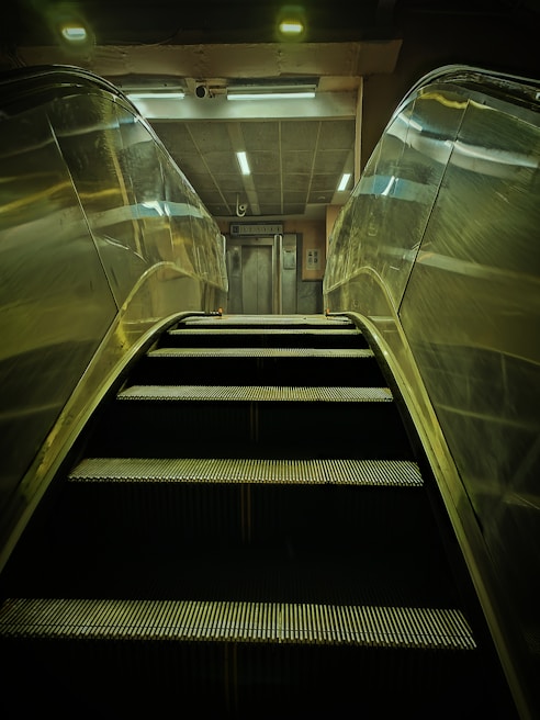 An escalator is ascending towards a well-lit area featuring an elevator. The scene is mostly metallic with reflections on the escalator sides, and overhead lighting provides a dim glow.
