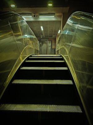 An escalator is ascending towards a well-lit area featuring an elevator. The scene is mostly metallic with reflections on the escalator sides, and overhead lighting provides a dim glow.
