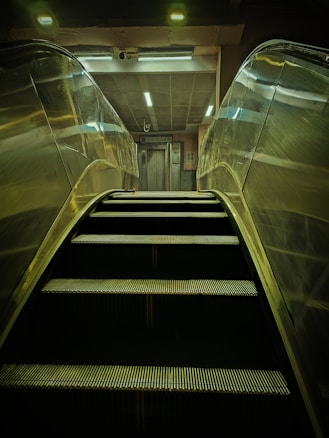 An escalator is ascending towards a well-lit area featuring an elevator. The scene is mostly metallic with reflections on the escalator sides, and overhead lighting provides a dim glow.