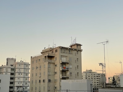 A collection of mid-rise apartment buildings is set under a clear sky during sunset. The buildings feature various antennas and satellite dishes on the rooftops. The architecture is plain and functional, typical of urban residential areas.