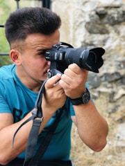 A person with short hair is holding a DSLR camera, looking through the viewfinder. They are wearing a blue t-shirt and a black wristwatch. The background shows a rough stone wall and some blurry natural elements, possibly grass.