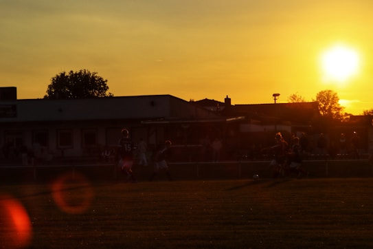 A sunset scene over a soccer field with players visible in silhouette, surrounded by a warm golden glow. The sun is low in the sky, casting long shadows and illuminating the sky with hues of orange and yellow. Trees and a building outline are in the background.