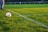 A soccer ball rests on a well-manicured grass field, bordered by fresh white lines marking the corner area. In the background, there are blurred figures and fencing, suggesting a sporting environment. One person's legs in jeans and sneakers are visible near the ball.