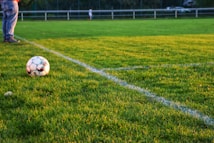 A soccer ball rests on a well-manicured grass field, bordered by fresh white lines marking the corner area. In the background, there are blurred figures and fencing, suggesting a sporting environment. One person's legs in jeans and sneakers are visible near the ball.