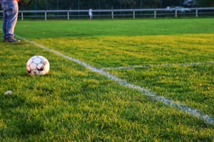 A soccer ball rests on a well-manicured grass field, bordered by fresh white lines marking the corner area. In the background, there are blurred figures and fencing, suggesting a sporting environment. One person's legs in jeans and sneakers are visible near the ball.