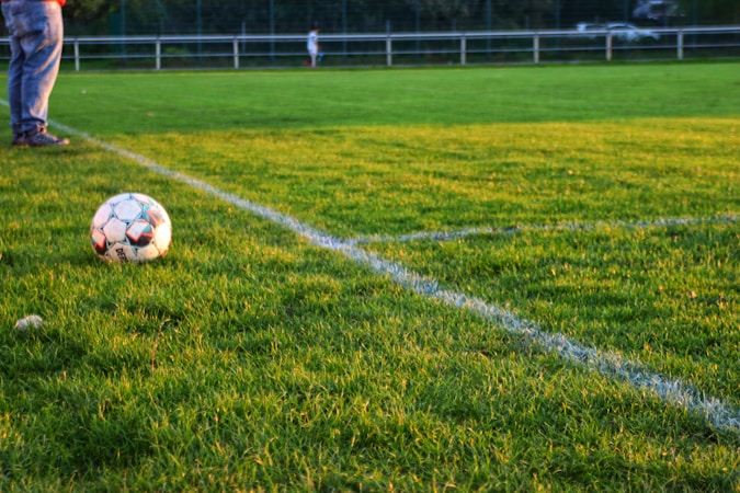 A soccer ball rests on a well-manicured grass field, bordered by fresh white lines marking the corner area. In the background, there are blurred figures and fencing, suggesting a sporting environment. One person's legs in jeans and sneakers are visible near the ball.
