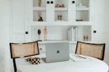 A cozy and minimalist workspace features a round white table with a modern laptop placed on it. Two woven chairs face the table, and nearby are a checkered mug and a notebook with pens. Behind, a white cabinet with shelves displays decorative items including books and small sculptures.