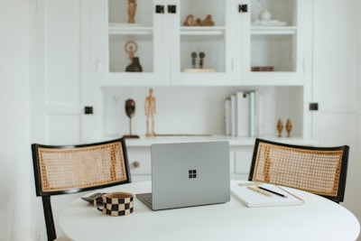 A cozy and minimalist workspace features a round white table with a modern laptop placed on it. Two woven chairs face the table, and nearby are a checkered mug and a notebook with pens. Behind, a white cabinet with shelves displays decorative items including books and small sculptures.