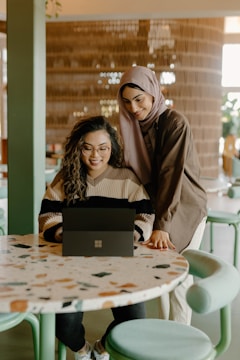 Two women are engaged in an activity at a table, one sitting and working on a laptop while the other stands next to her, both smiling and appearing friendly.