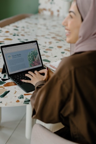 A person wearing a headscarf is sitting at a table and using a laptop. The screen displays a document titled 'Coral Reefs Saving our Oceans'. The table has a mosaic design with various colors.