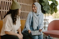 Two women are sitting together, smiling and engaging in conversation. One woman is wearing a headscarf and holding a tablet with a stylus, while the other has curly hair and a knit hat. They are seated on comfortable chairs in a warmly lit room with plants and patterned walls.