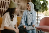 Two women are sitting together, smiling and engaging in conversation. One woman is wearing a headscarf and holding a tablet with a stylus, while the other has curly hair and a knit hat. They are seated on comfortable chairs in a warmly lit room with plants and patterned walls.