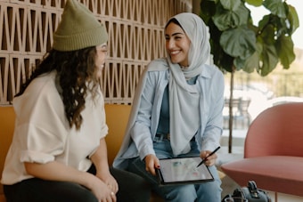 Two women are sitting together, smiling and engaging in conversation. One woman is wearing a headscarf and holding a tablet with a stylus, while the other has curly hair and a knit hat. They are seated on comfortable chairs in a warmly lit room with plants and patterned walls.