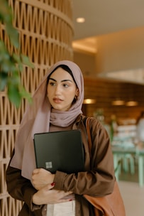 A woman wearing a light purple hijab and a brown shirt is holding a laptop. She has a brown shoulder bag and is standing indoors with a patterned brown wall and some greenery to her left. The background features a modern, softly lit room with furniture and decor.