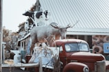 A retro red truck is parked in front of a building with a large metal roof. On the truck's bed, there is a large sculpture of a moose and several cow figures mounted on the roof of the adjoining structure. The scene includes some autumnal foliage and scattered pieces of vegetation in the foreground.