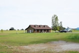 A rustic wooden house is situated in the middle of a vast green lawn with a few trees scattered around. A pickup truck is parked nearby on a gravel driveway, and a small trailer is visible behind it. In the background, expansive open fields stretch out under a pale sky.