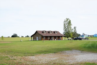 A rustic wooden house is situated in the middle of a vast green lawn with a few trees scattered around. A pickup truck is parked nearby on a gravel driveway, and a small trailer is visible behind it. In the background, expansive open fields stretch out under a pale sky.