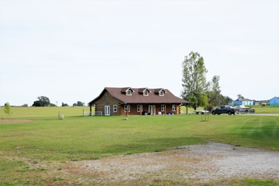 A rustic wooden house is situated in the middle of a vast green lawn with a few trees scattered around. A pickup truck is parked nearby on a gravel driveway, and a small trailer is visible behind it. In the background, expansive open fields stretch out under a pale sky.